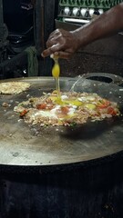 A street food vendor in Madurai cracks fresh eggs over a sizzling mix of onions, tomatoes, and spices on a giant hot griddle, preparing a signature egg dish right in front of hungry customers.