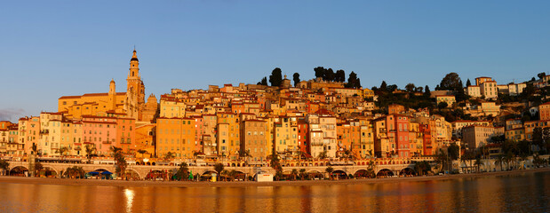 Panoramic sunset view of Menton, France, with colorful old town buildings glowing in warm light, set against the Mediterranean Sea and blue sky on the French Riviera