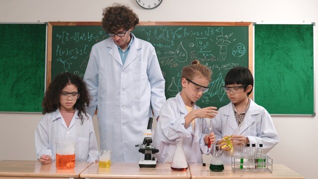 Boy pour solution in beaker while american girl inspect colored liquid. Energetic student support his friend doiing experiment while teacher looking children explore and discover innovation. Pedagogy.