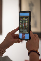 A woman holds a phone with a camera app and takes pictures of oil paintings featuring Christian saints.
