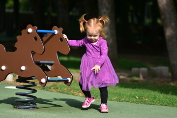 Red-haired toddler girl in purple dress and pink shoes walking near brown horse spring rider at...