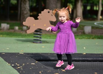 Red-haired toddler girl in purple dress and pink sandals standing on playground trampoline, smiling...