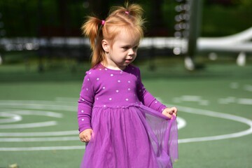 Fototapeta premium Red-haired toddler girl in long-sleeve purple polka dot dress holding skirt while standing in playground, thoughtful expression, green rubber surface, soft sunlight, outdoor portrait.