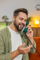 Smiling young man making wired telephone call conversation with friends sitting on couch at home....