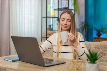 Caucasian woman freelancer types on laptop sitting at home table, holding cup of coffee or tea,...
