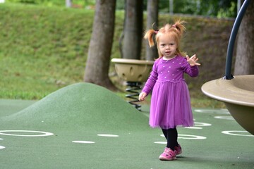 Red-haired toddler girl in purple dress and pink sandals walking on green playground surface,...