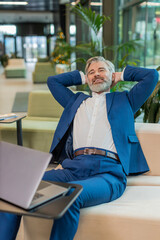 Happy businessman sitting comfortably with hand behind head in modern office lobby closing laptop...