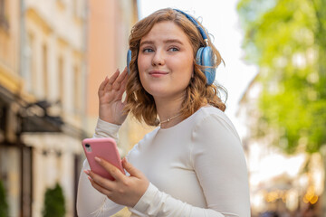 Happy relaxed overjoyed redhead young woman in wireless headphones listening energetic disco music in smartphone app dancing outdoors. Red hair girl walking in urban city street. Town lifestyles.