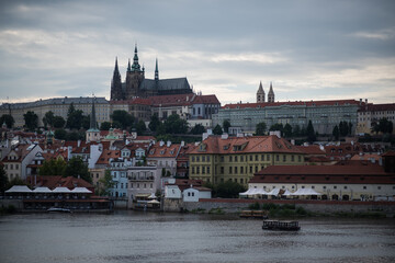 Obraz premium Prague Castle complex and the city's historic skyline, as seen from across the Vltava River. In the foreground, a row of charming, colorful buildings with traditional red-tiled roofs lines 