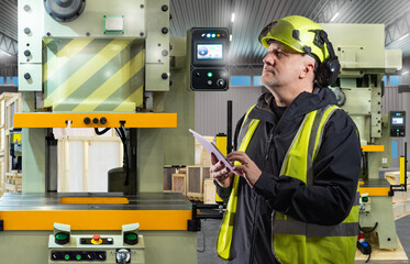 Factory worker in safety gear using digital tablet to monitor and control industrial press machine,...