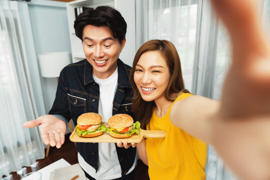Smiling host chef influencers taking photo with smartphone by selfie straight view holding double hamburger on cooking show special easy dish homemade on social media channel live streaming. Infobahn.