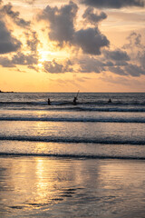 Distant silhouettes of three fishermen wading into the Arabian Sea at Caranzalem Beach, Goa, on a monsoon evening, with golden sunset, dramatic clouds, and shimmering water reflections.