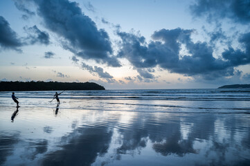 Dusk at Caranzalem Beach, Goa, with dramatic cloudy skies, waves, and two backlit cliffs. Two...