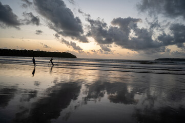 Obraz premium Dusk at Caranzalem Beach, Goa, with dramatic cloudy skies, waves, and two backlit cliffs. Two fishermen silhouettes cast their lines, their reflections mirrored in the wet sand and sea.