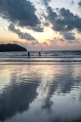 Two fishermen cast their nets in the Arabian Sea from Miramar Beach, Panjim, Goa, as the sun sets on the horizon, with a backlit cliff, dramatic monsoon clouds, and an orange glow in the evening sky.