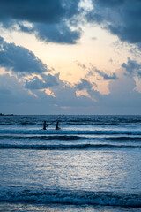 Fototapeta premium Two fishermen silhouetted at dusk cast their nets in the Arabian Sea from Siridao Beach, Panjim, Goa, beneath a monsoon sky painted orange with clouds over the calm evening waves.