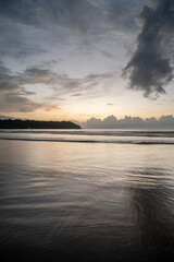 Obraz premium Landscape view of Miramar Beach Panjim Goa during monsoon sunset, with Arabian Sea waves, dramatic grey clouds, yellow-orange horizon glow, backlit cliffs, wet sand textures, and shimmering reflection