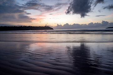 Obraz premium Landscape view of Caranzalem Beach in Goa, India, during monsoon sunset with sand textures in foreground, backlit cliff, waves in Arabian Sea, dramatic clouds, and warm yellow-orange sky reflection.