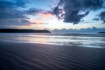Fototapeta premium Landscape shot of Miramar Beach, Goa, India, after sunset at dusk with wet sand line textures, waves, two backlit cliffs, cloud line at horizon, and surreal sky with yellow, pink, orange, and dramatic
