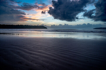 Obraz premium Landscape shot of Miramar Beach, Goa, India, after sunset at dusk with wet sand line textures, waves, two backlit cliffs, cloud line at horizon, and surreal sky with yellow, pink, orange, and dramatic