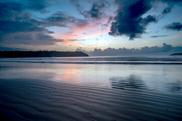 Obraz premium Landscape shot of Miramar Beach, Goa, India, after sunset at dusk with wet sand line textures, waves, two backlit cliffs, cloud line at horizon, and surreal sky with yellow, pink, orange, and dramatic