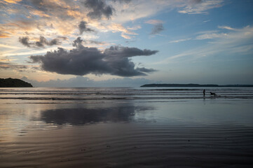 Landscape of Miramar Beach Goa after sunset at dusk, wet sand textures with footprints, waves, backlit cliffs, silhouette of girl and dog playing, sky in yellow, pink, orange, blue, grey clouds