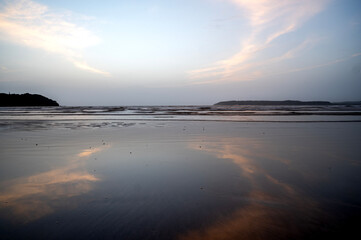 Obraz premium A shot of heart-shaped cirrus white clouds in the light blue sky at Miramar Beach, Goa, reflected in the water, with gentle waves and two distant cliffs in the background.