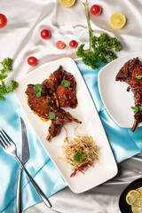 Top view of lamb chops on white plates with fresh salad, lemon slices, cherry tomatoes, coriander leaves, fork and knife on white and blue satin background.
