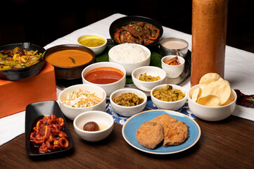 Goan fish thali with rawa fried kingfish, crab, calamari, prawns, fish curry, rice, papad, dal, thrissur, sol curry, gulab jamun, served in bowls and plate with copper water bottle on brown table