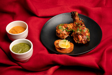 Juicy chicken tangdi kebab with fresh salad in black plate on red table mat background, served with green chutney and yellow sauce in small white bowls