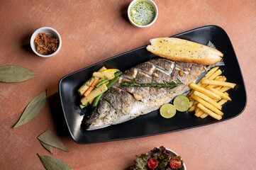 Top view of pan-fried seabass with Romesco sauce, sautéed broccoli, wilted bok choy, almond flakes, bread, lemon slices, and fries on a black plate, styled with butter sauce and garnish bowls.