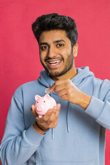 Happy Indian young man holding piggy bank, saving money by dropping coins, planning future expenses...