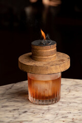 Smoked Old-Fashioned cocktail in highball glass with large ice ball on marble table, cocktail...