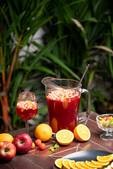 Fototapeta premium Sangria served in a glass jug and wine glass on a table, with whole and sliced fruits like oranges and apples in a plate, with blurred green plants in the background.