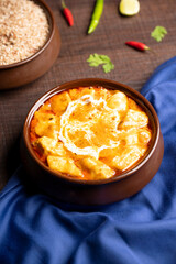 A top shot of Paneer Butter Masala, cottage cheese slow-cooked in rich buttery tomato and cashew nut gravy, served in a rustic brown pot on blue satin with green and red chilies.