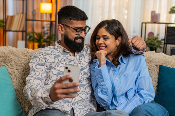 Indian couple man and woman sitting on home sofa, celebrating lottery win on smartphone with emotions of surprise and joy. Young family looks excited and happy while reacting to jackpot result indoors