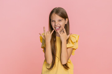 Young child girl dances joyfully and makes rock-and-roll gesture with raised hand, expressing playful energy confident mood. School kid on pink background, feeling cool enthusiastic during performance