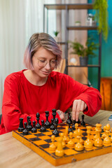 Young woman in glasses focuses on chess move at home living room over smart wooden board plotting...