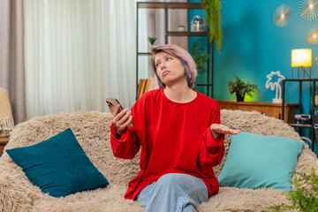 Young woman sadly looking at smartphone screen at home, upset by disappointing message, shaking...