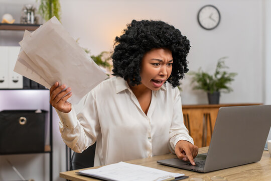 African American businesswoman at home office sees laptop crash glitch panics tossing papers urgently. Freelancer girl at table hits power button reboot hoping data safe returns and virus cleared soon