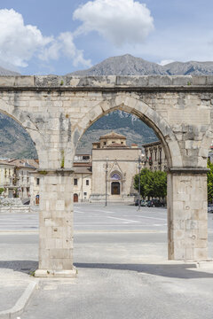 View of ancient stone archway frames a sun-drenched piazza with a domed building, backed by rugged mountains against a bright sky, Sulmona, Abruzzo, Italy.