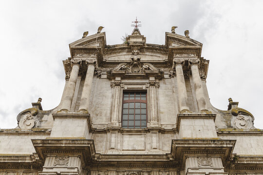 View of a marble facade with a striking window and ornate carvings reaching up to a cross against a cloudy sky, Sulmona, Abruzzo, Italy.