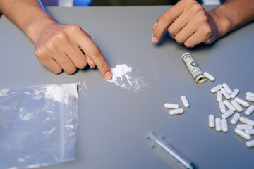 Drug addict male preparing dose of cocaine on gray table, surrounded by syringe, pills and...