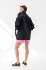 Fototapeta premium Elegant woman in black windbreaker jacket and pink shorts on light background. Fashionable female style. Studio photography. Vertical photo. High quality photo