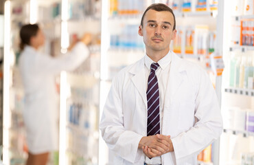 At pharmacy, smiling man specialist stands in hall with open display and gestures to invite...