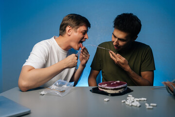Two friends enjoying drug-fueled party licking icing dripping from cake, with pills and alcohol...