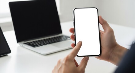 A person using a smartphone with a blank white screen, with a laptop in the background (mockup).