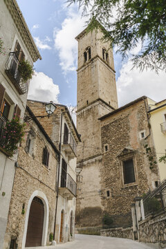 View of an old stone church tower rising high above the narrow street with aged stone buildings under a partly cloudy sky, Pacentro, Abruzzo, Italy.