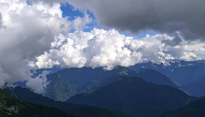 Fototapeta premium Fluffy white clouds drift over a range of dark, tree-covered mountains, with glimpses of snow-capped peaks in the distance.