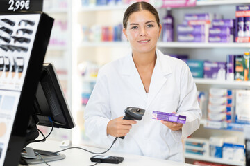 Girl pharmacist in cash area of pharmacy works with computer and goods, scans drugs, checks availability. Chemists shop has wide selection of cosmetics, care products, medicines